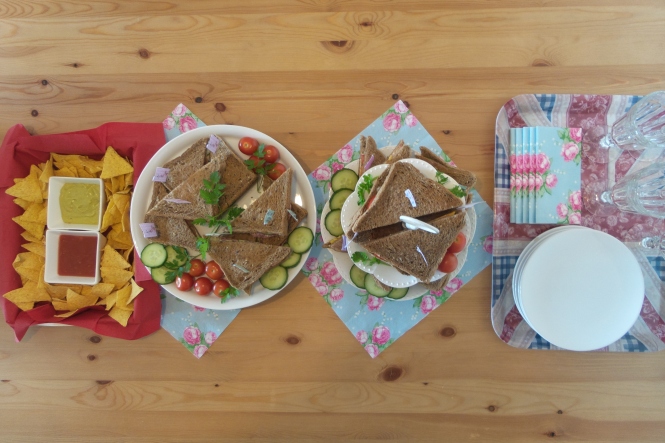 Von links auf einem Tisch: Tortilla Chips mit Dip und zwei Platten mit Sandwiches. Ganz rechts stehen Teller, Getränke und Servietten
